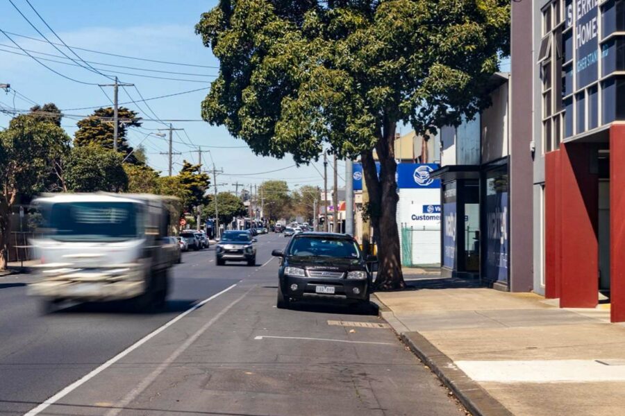 Packington Street in Geelong - vehicles moving and parked along the street; shops and trees on the side; powerlines and blue sky above