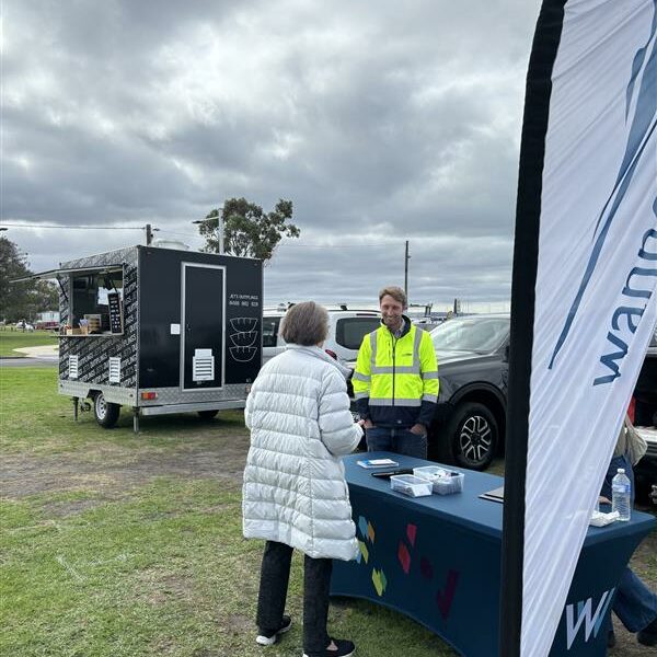 An outdoor pop-up event with Wannon Water flag and table. A Wannon Water employee is chatting to a community member wearing a light grey puffa coat. Coffee cart, park cars and trees in background; grey clouds above.