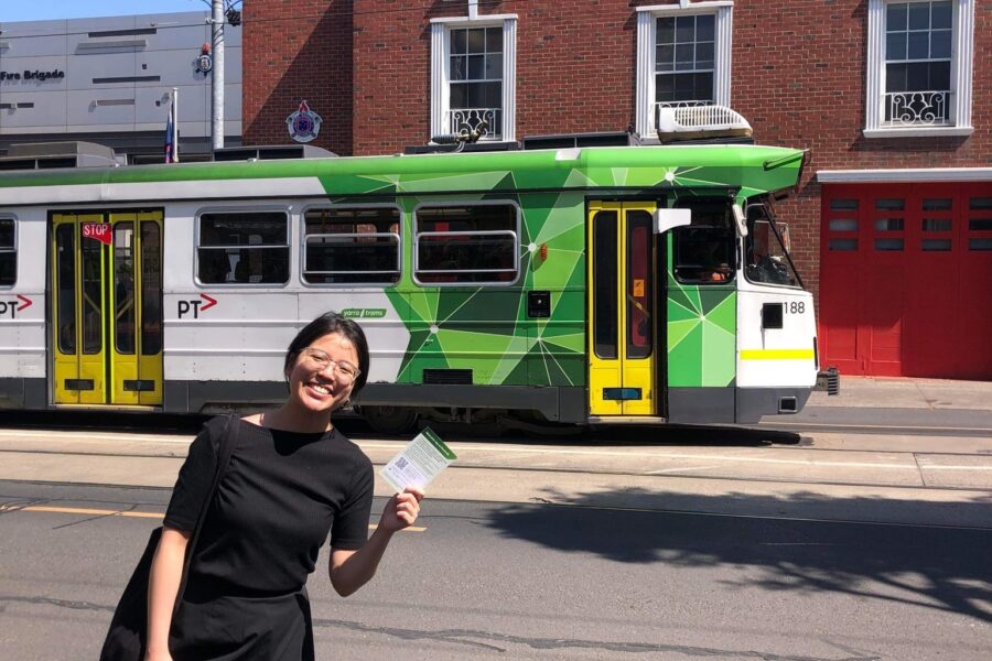 Capire consultant Charlotte stands in front of a tram holding a postcard for the survey. Charlotte is wearing a black top and long black skirt; she has short dark hair and is smiling. There is a red-brick building in the background.