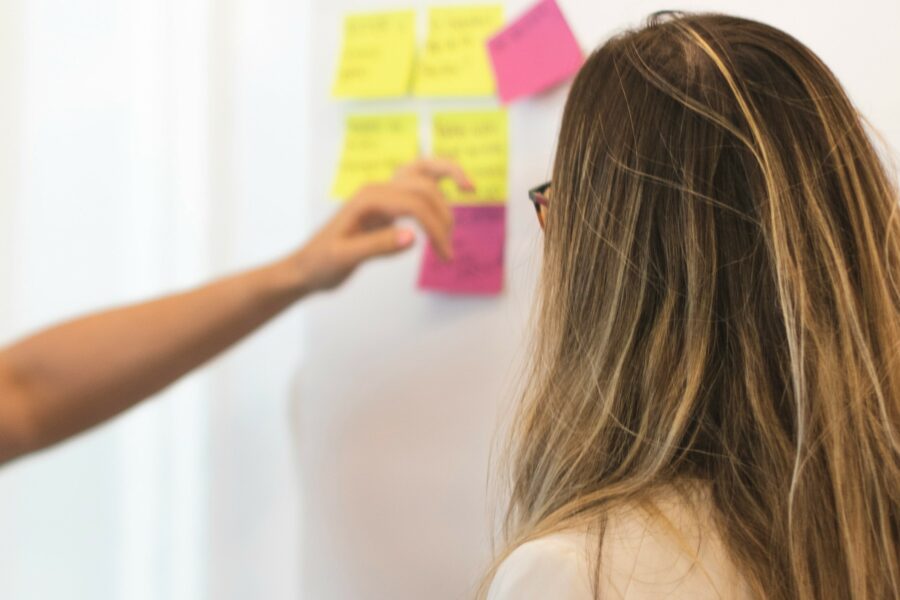 A woman standing, facing a white board covered in yellow and pink Post-it notes. She is facing the board, so only the back of her head is visible. She wears a white shirt and has long straight blonde-brown hair.