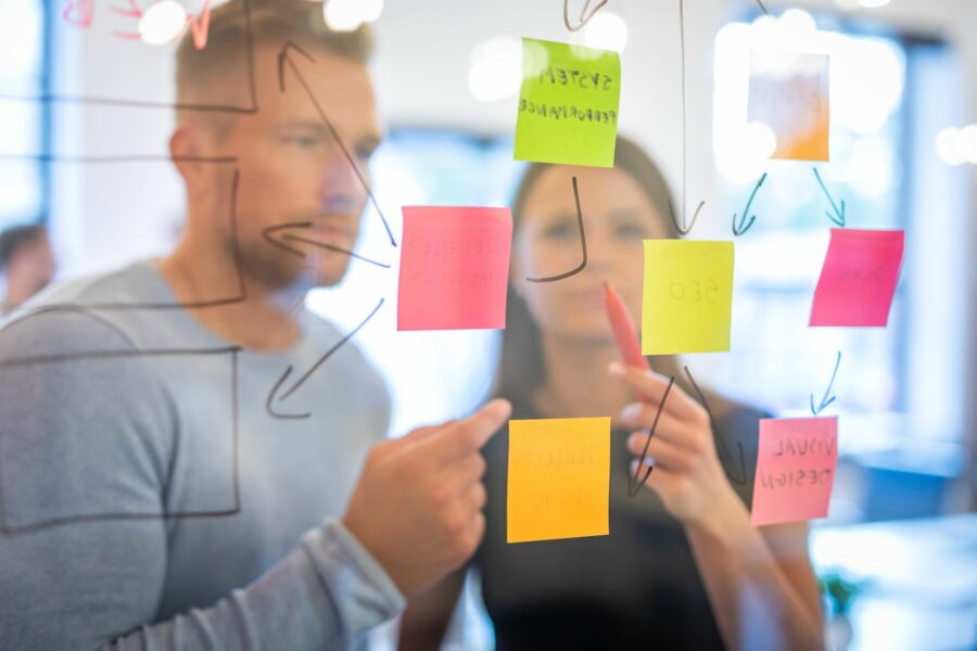 A deliberate engagement session. Two people, a man and a woman, stand behind a clear screen covered in colourful Post-it notes.