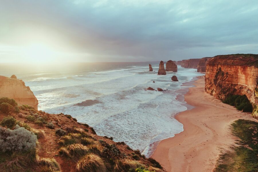 The Twelve Apostles: several limestone stacks standing in the shore of Victoria's southern coastline. The ocean, sand and surrounding cliffs are all visible. Sun filters through the sky above.