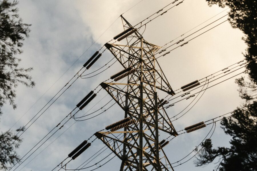 A transmission tower and power lines, viewed from below. Sky in background; trees on either side.
