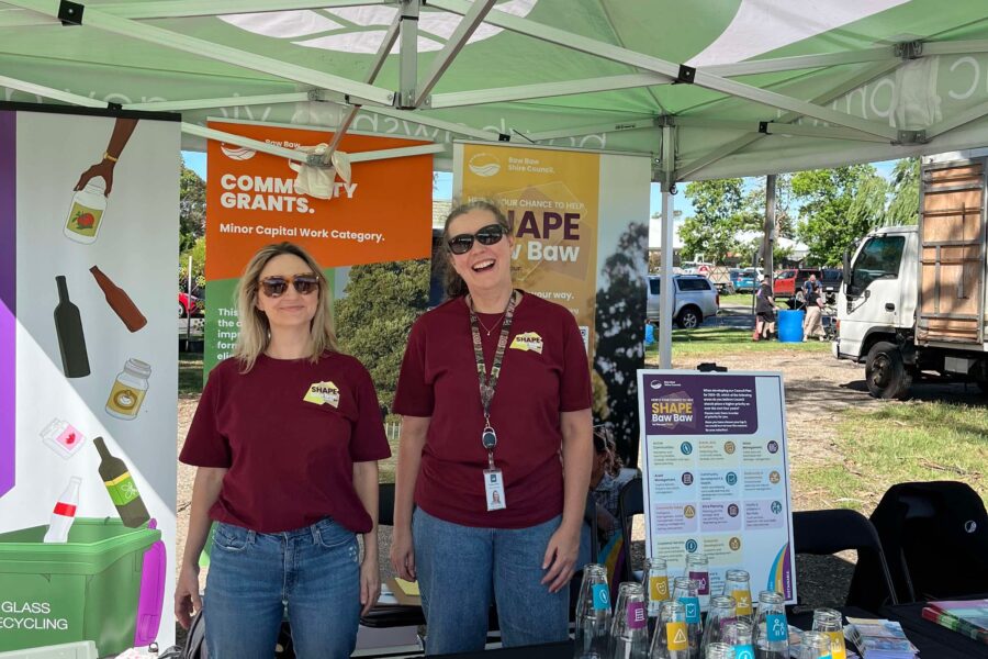 Pop-up event at an outdoor market. Two community engagement consultants stand behind a desk displaying items for engagement activities: posters, pens, Post-it notes, bottles, tokens. They are both smiling and wearing maroon 'Shape Baw Baw' t-shirts, jeans and sunglasses.