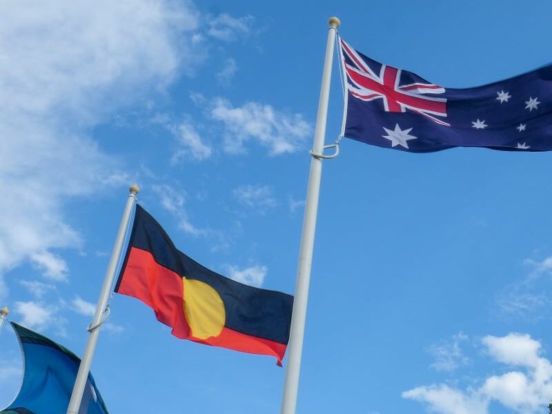 The Torres Strait Islander flag, Aboriginal flag, and Aboriginal flag fluttering against a blue sky.