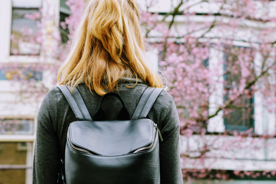 Back of a female student, wearing a black backpack, walking towards a school building.