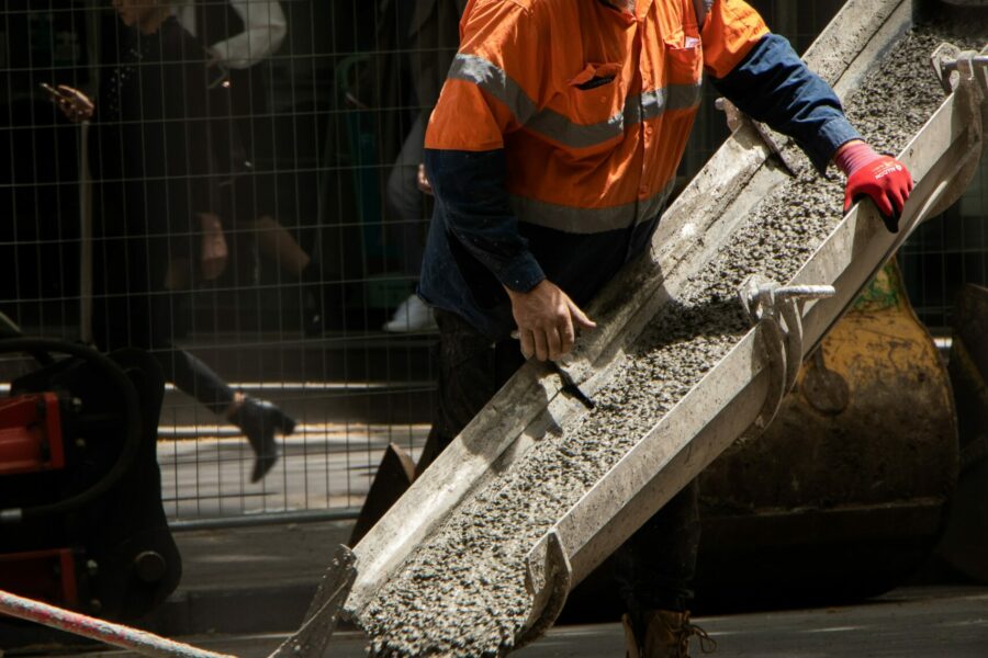A male construction worker wearing a white hat and orange work shirt is pouring concrete.