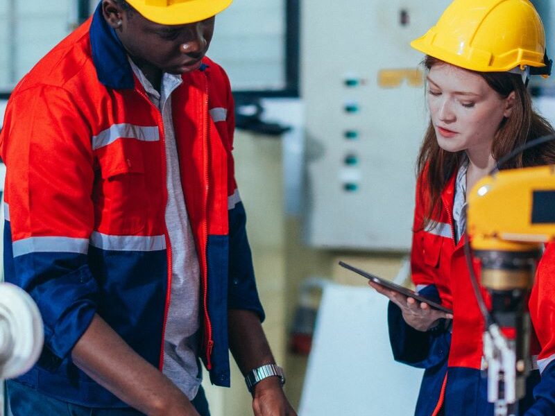 Students wearing yellow hard hats and hi-vis shirts looking at a blueprint. Manufacturing equipment in background.