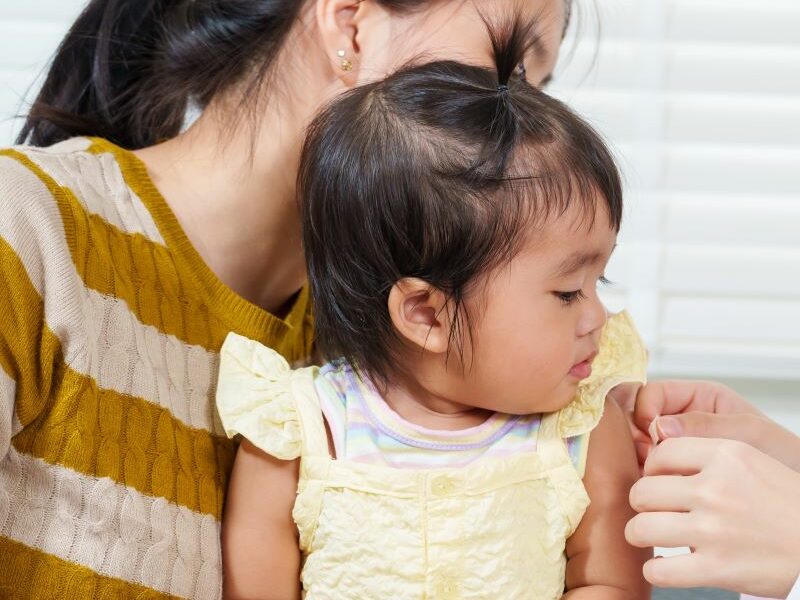 An Asian toddler sits on her mother's lap, while a female doctor applies a Band-Aid to her arm following vaccination.