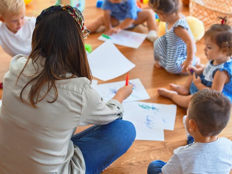A teacher and group of toddlers sit in a circle, drawing.