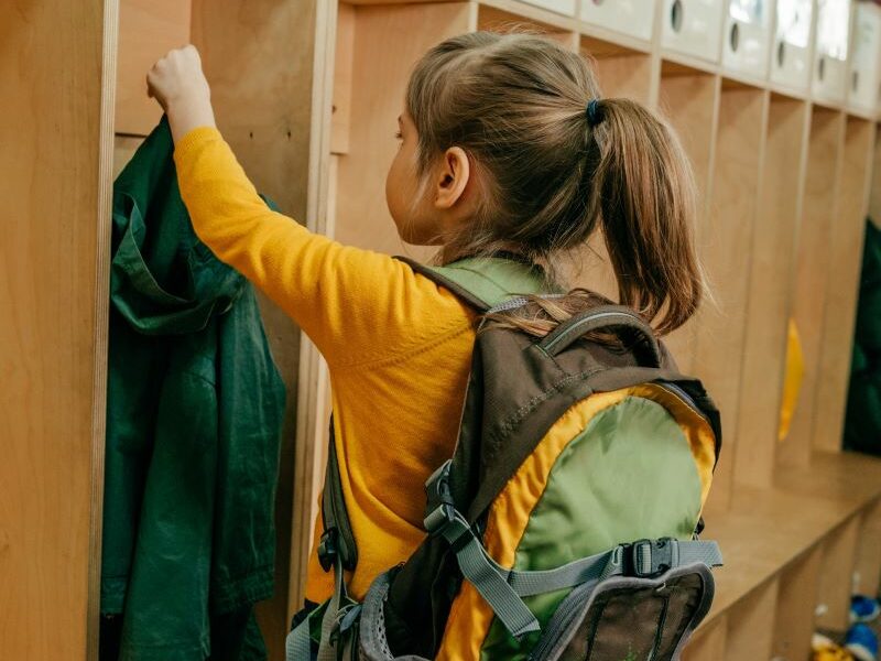 A young girl attending kindergarten. She is wearing a backpack and hanging her coat in her locker.