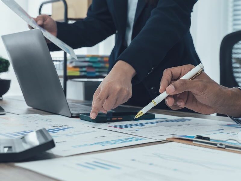 Three people, dressed in business attire, sit or stand around an office desk. Laptops, print-outs and a calculator sit neatly on top of the desk.