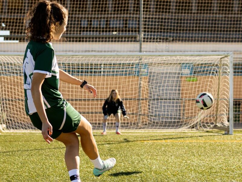 A girl taking a penalty kick on a grass soccer field. Goalie and soccer ball in background