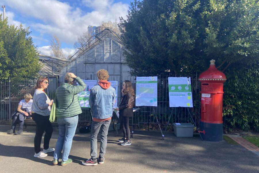 A community pop-up event: a small group of people are gathered outside, around some boards featuring information about neighbourhood batteries. A council representative stands in front of one of the boards