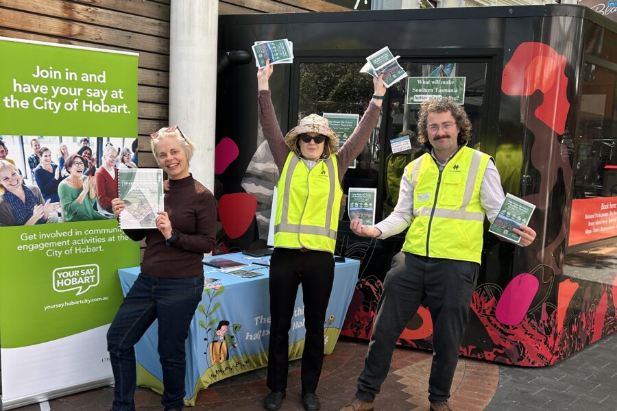 Three Council workers, wearing hi-vis yellow vests, standing in front of a pop-up stall for the City of Hobart. They are smiling and posing with a report and leaflets. They are standing in front of a table and 'Your Say Hobart' banner