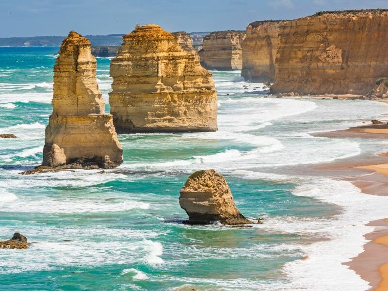 The Twelve Apostles, Port Campbell National Park. A group of limestone rock stacks off the coast. Orange sand, aqua-blue water, and bright blue sky