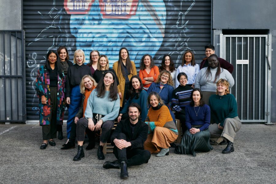 The Capire team, a diverse group of 20 staff members, wearing colourful clothes, standing/sitting in an outdoor urban environment