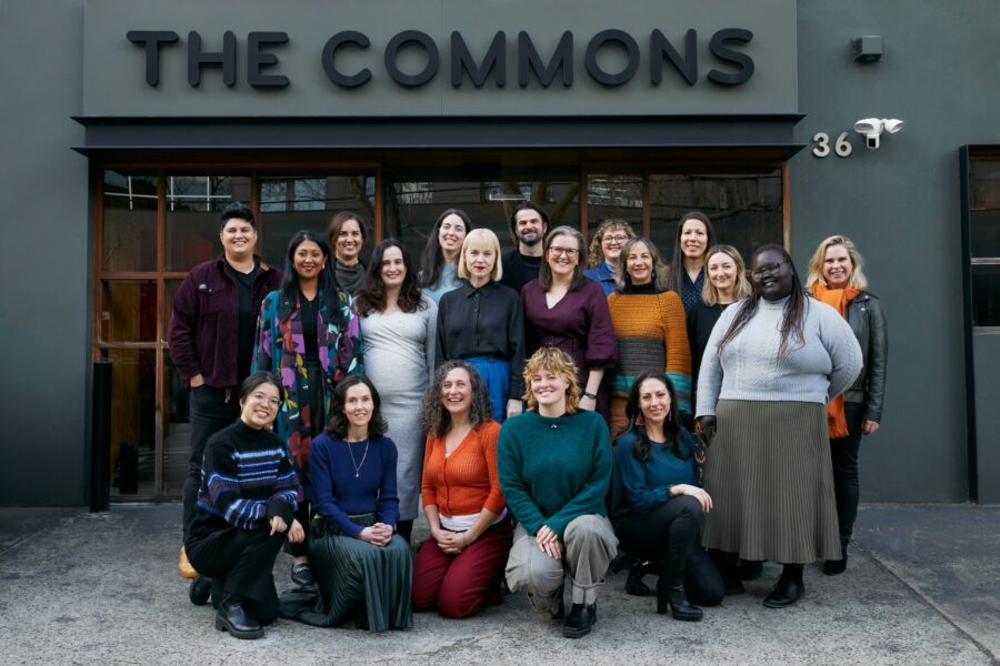 The Capire team, a group of 19 diverse people, stand/sit for a group photo outside of the Commons. They are smiling and dressed in colourful winter clothes.