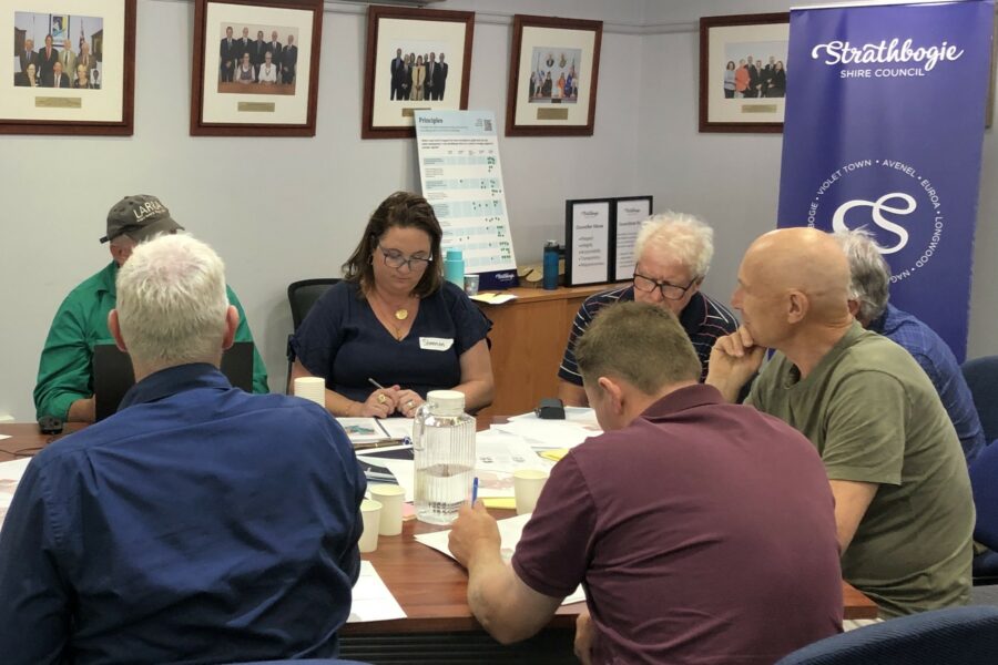 A group of people, sitting around a table, deliberating. They are in a Council room with framed pictures hanging on the wall.
