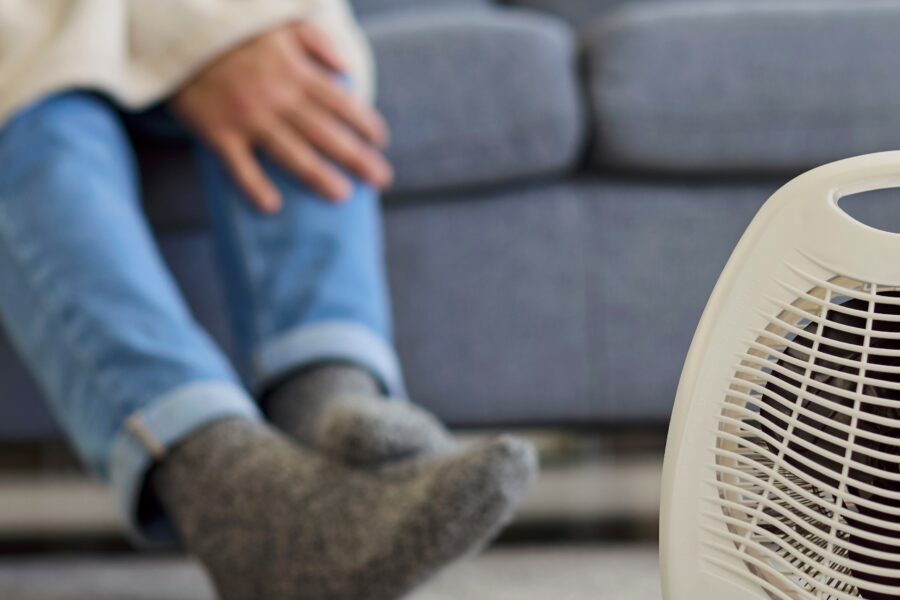 A person sitting on a couch with a blanket, wearing jeans and slippers; small electric heater in foreground. Only the person's legs and hand are visible