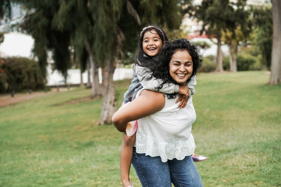 A mother carrying her daughter on her back in a public park. They are both smiling and laughing.